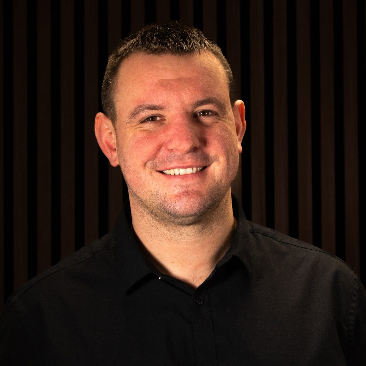 Head‑and‑shoulders portrait of Sam Hewitt, a young man with short brown hair and a smile, wearing a dark collared shirt. The background is a softly lit dark-toned setting