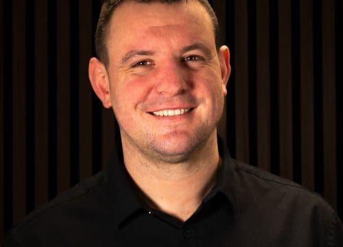 Head‑and‑shoulders portrait of Sam Hewitt, a young man with short brown hair and a smile, wearing a dark collared shirt. The background is a softly lit dark-toned setting
